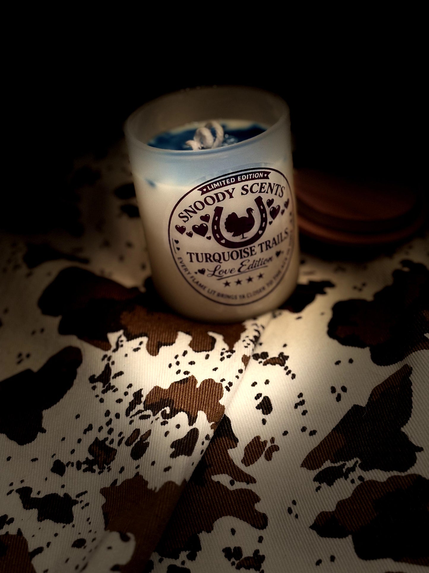 Turquoise Trails Love Edition candle glowing in low light on cowhide print fabric, wood lid set behind it