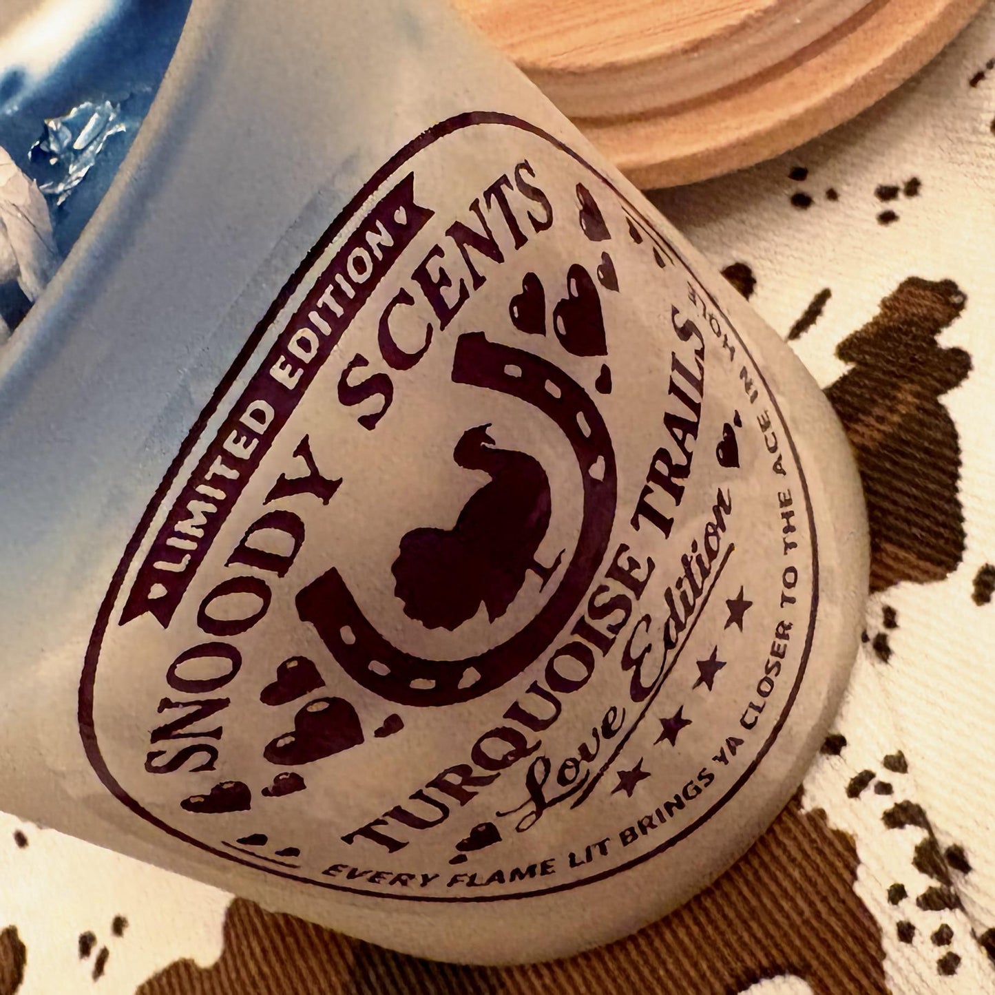 Tilted Snoody Scents Turquoise Trails Love Edition candle showing horseshoe logo label and blue wax inside