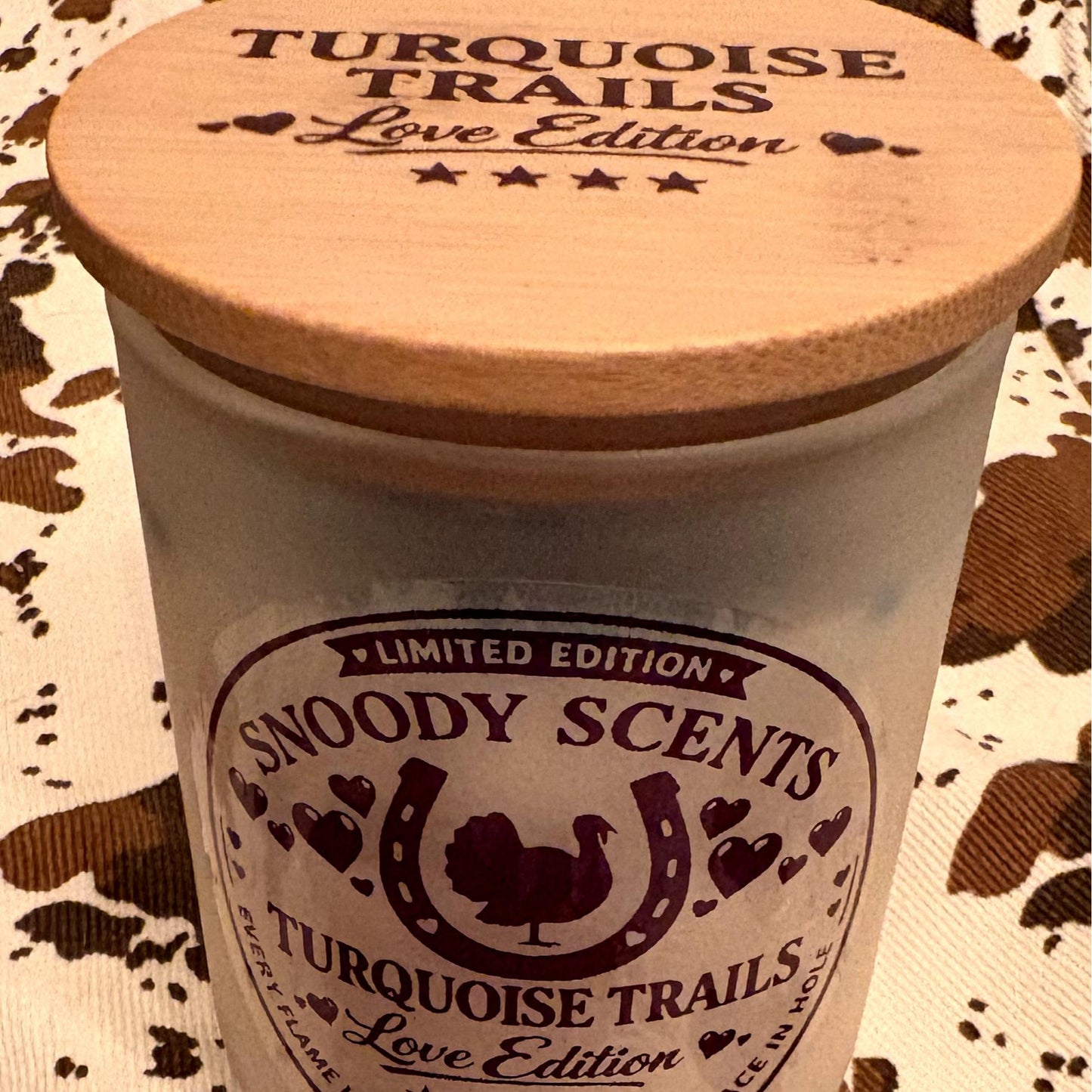 Close-up of the Snoody Scents Turquoise Trails Love Edition candle label and wood lid on cowhide print fabric